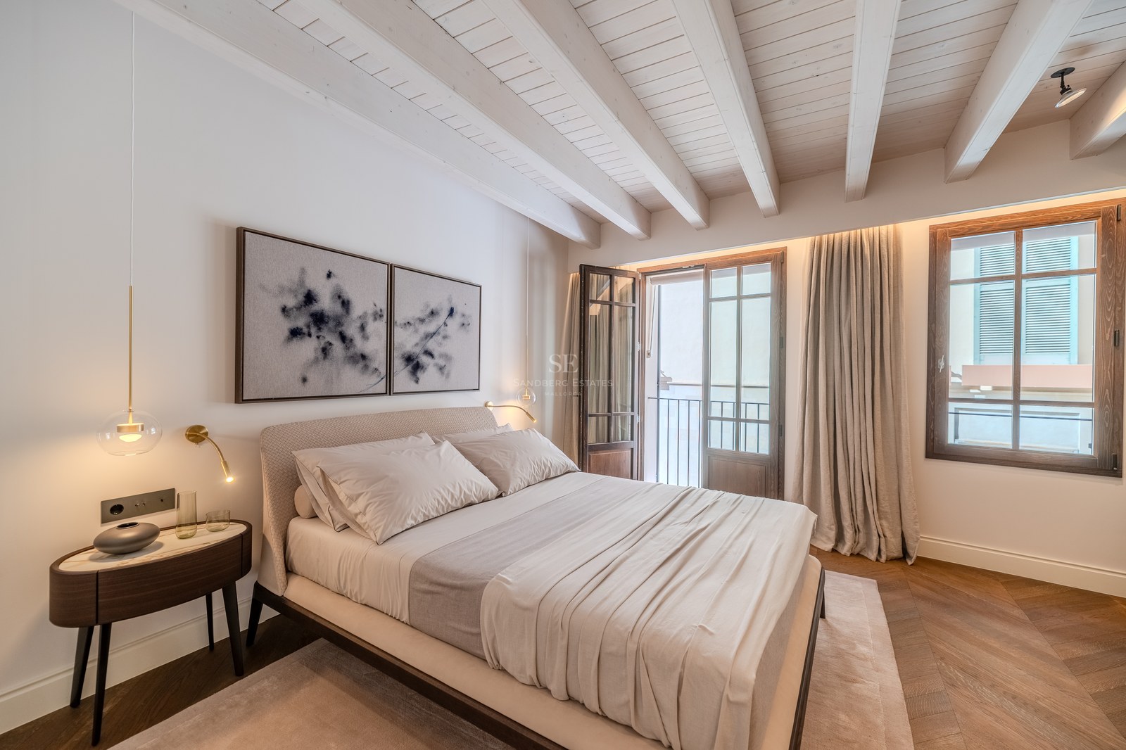 Chambre de luxe avec plafond à poutres blanches, parquet en chevrons, porte-fenêtre et décor neutre.