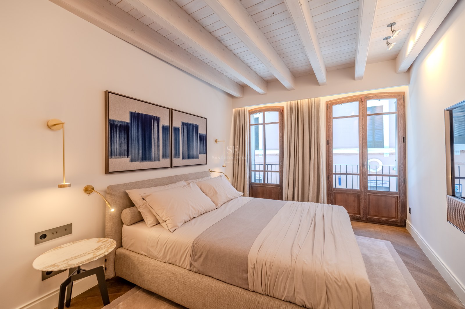 Chambre de luxe avec plafonds à poutres blanches, portes-fenêtres en bois et appliques en laiton.