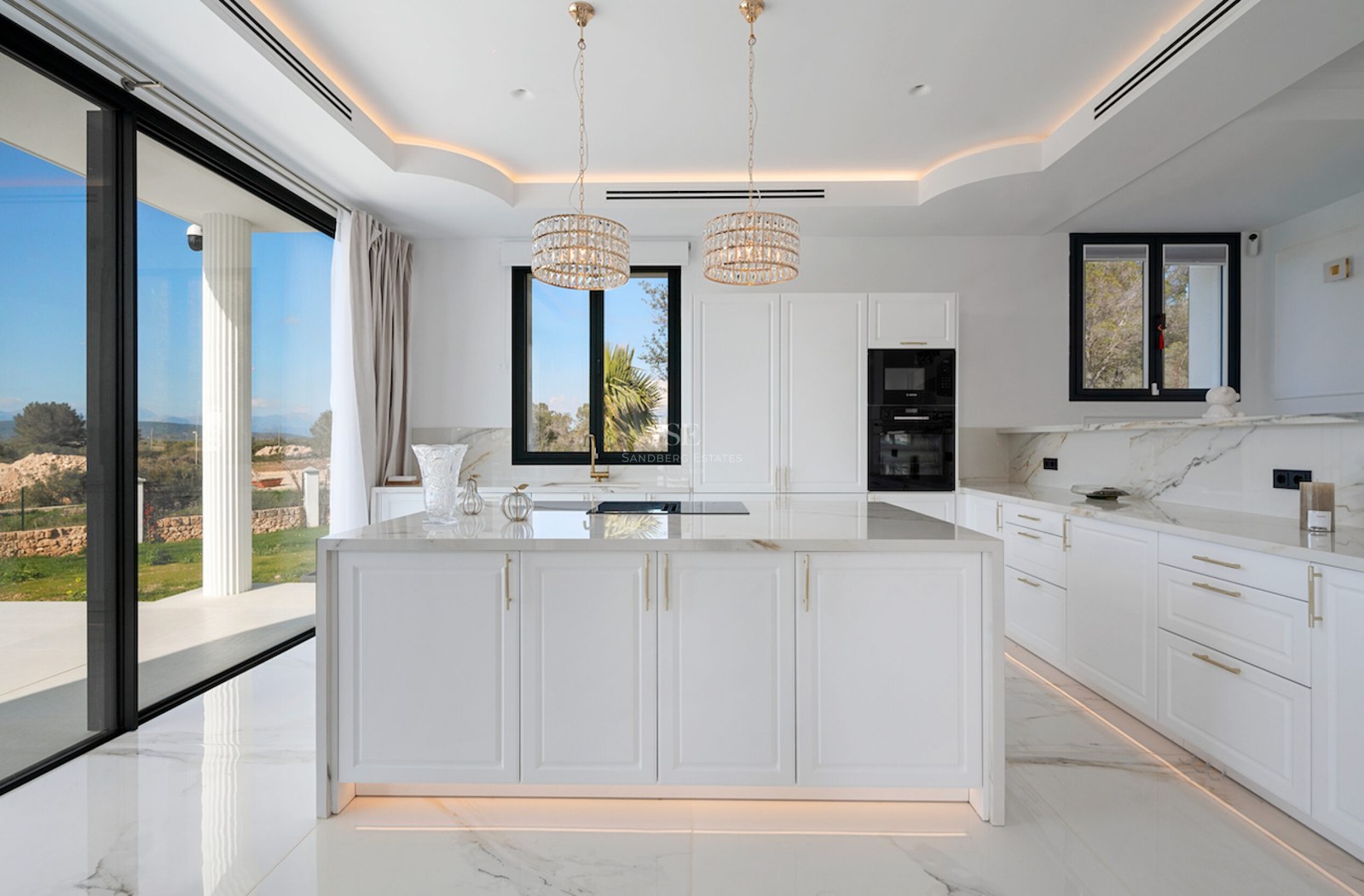 Cocina moderna y luminosa con gran isla de mármol, detalles dorados y ventanales de suelo a techo hacia el jardín.