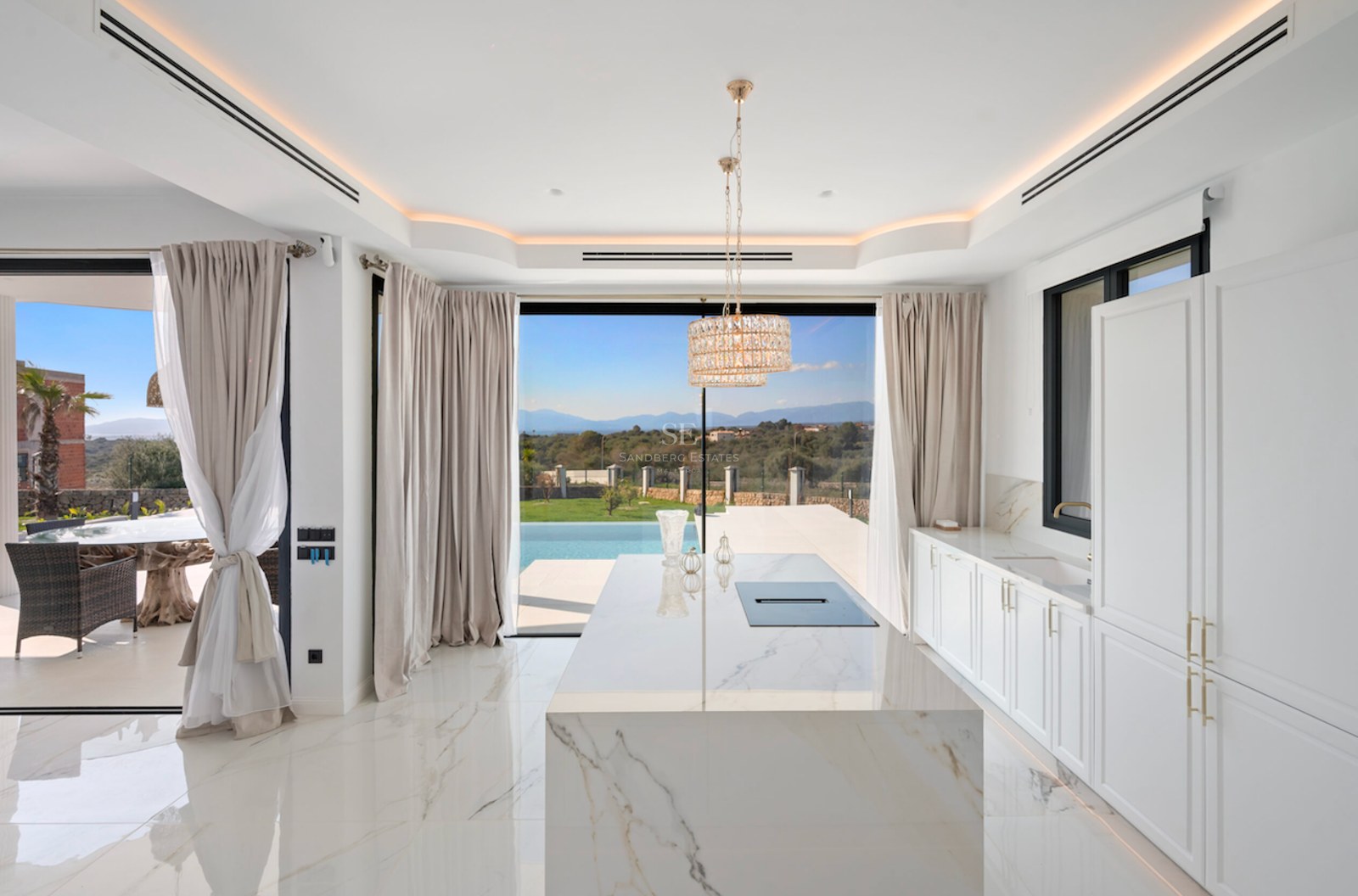 Cocina moderna con gran isla de mármol blanco, muebles blancos y grandes ventanales con vistas a la piscina y el paisaje.