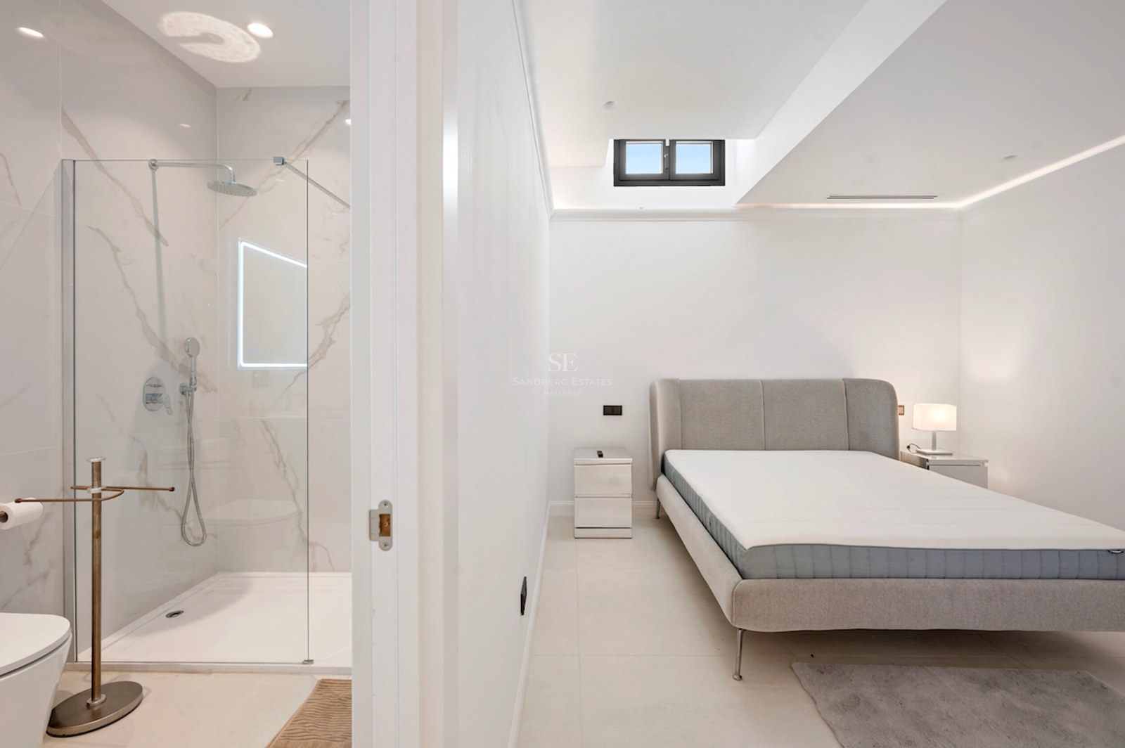 Dormitorio moderno con cama gris y puerta abierta hacia una ducha de mármol con mampara de cristal.