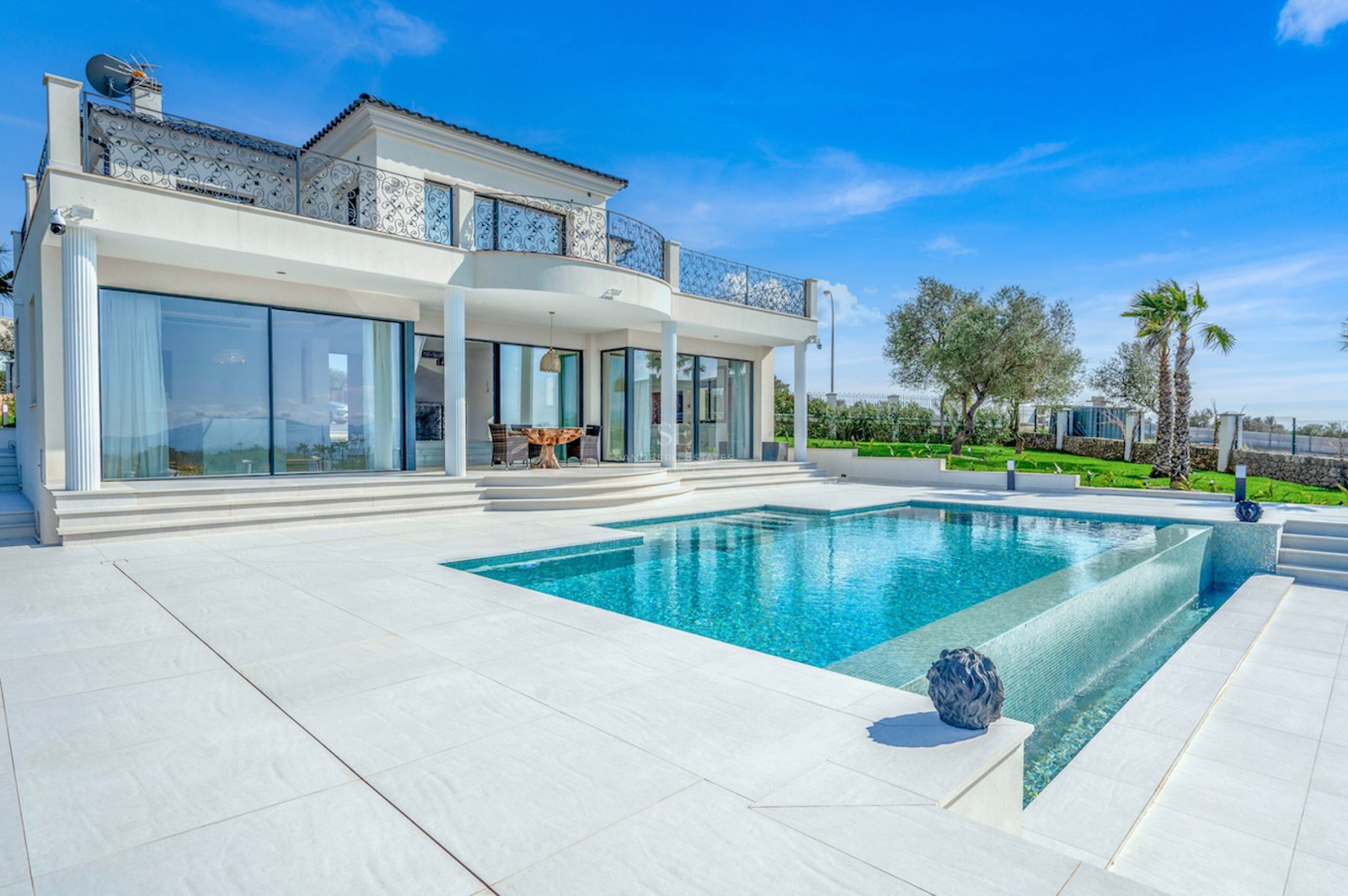 Villa blanca de estilo mediterráneo moderno con piscina infinita, terraza de piedra clara y jardín bajo cielo azul.