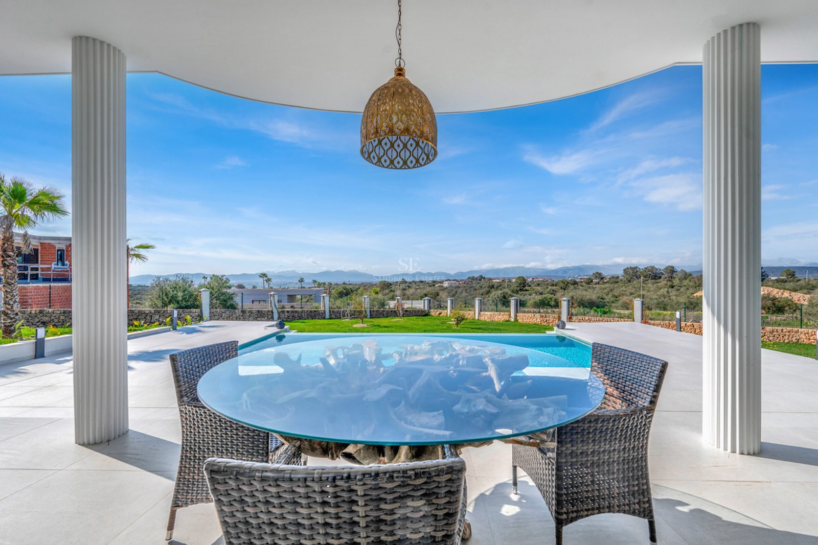 Terraza cubierta con mesa de cristal y sillas de mimbre frente a una piscina con vistas a la montaña.