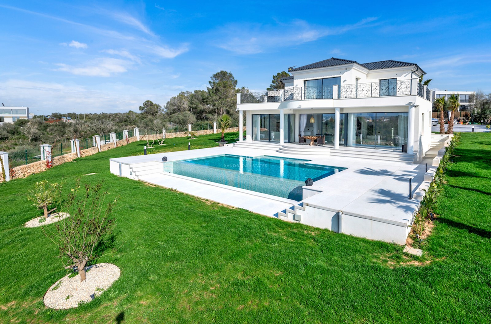 Vista de una villa blanca de dos plantas con piscina integrada, terraza de piedra y amplio jardín bajo cielo azul.