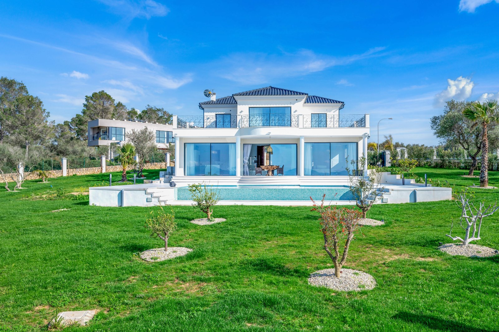 Fachada frontal de una villa de lujo blanca con piscina y amplio jardín bajo un cielo despejado.