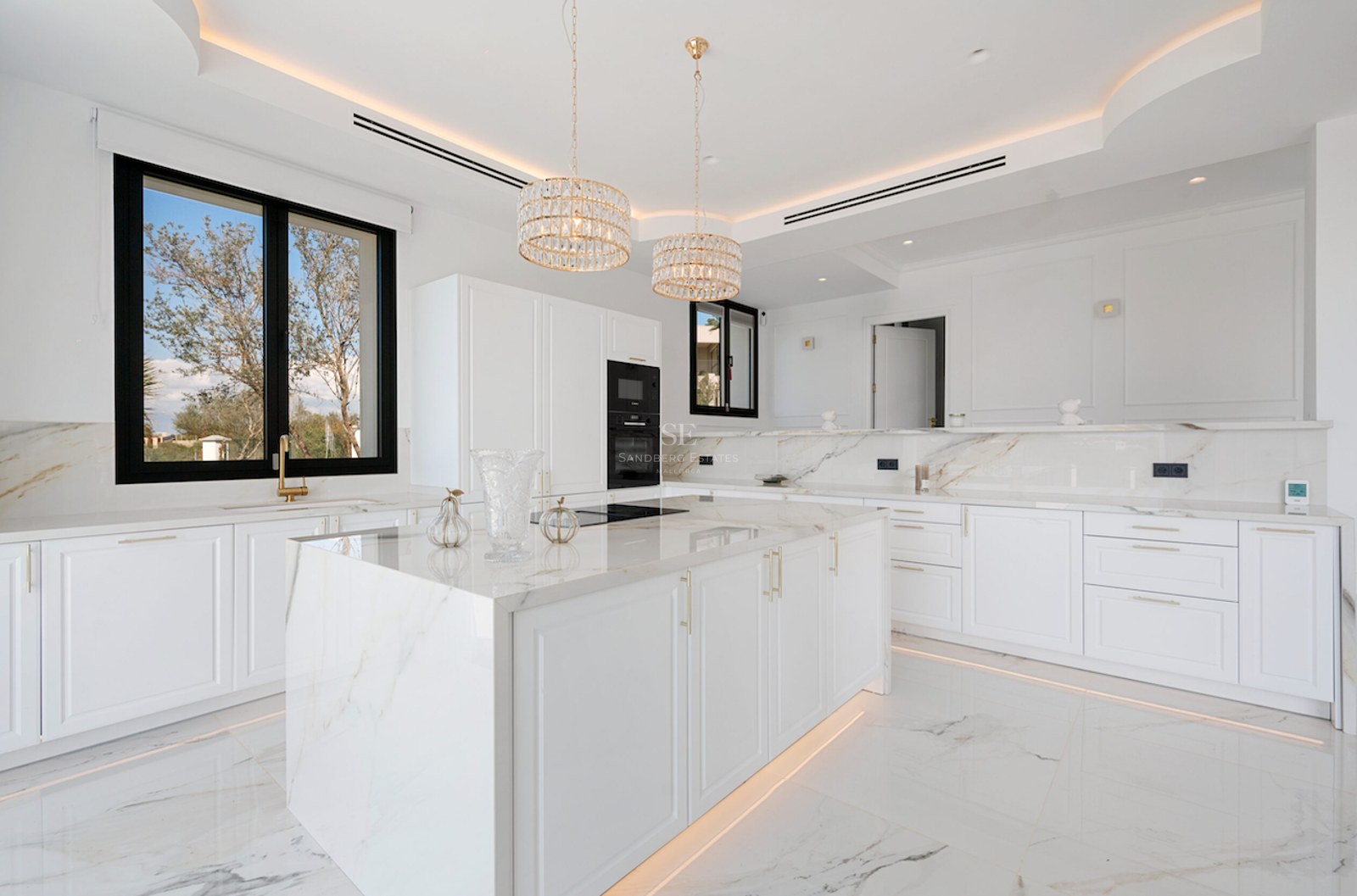 Cocina moderna y luminosa con isla central de mármol, detalles dorados y amplios ventanales.