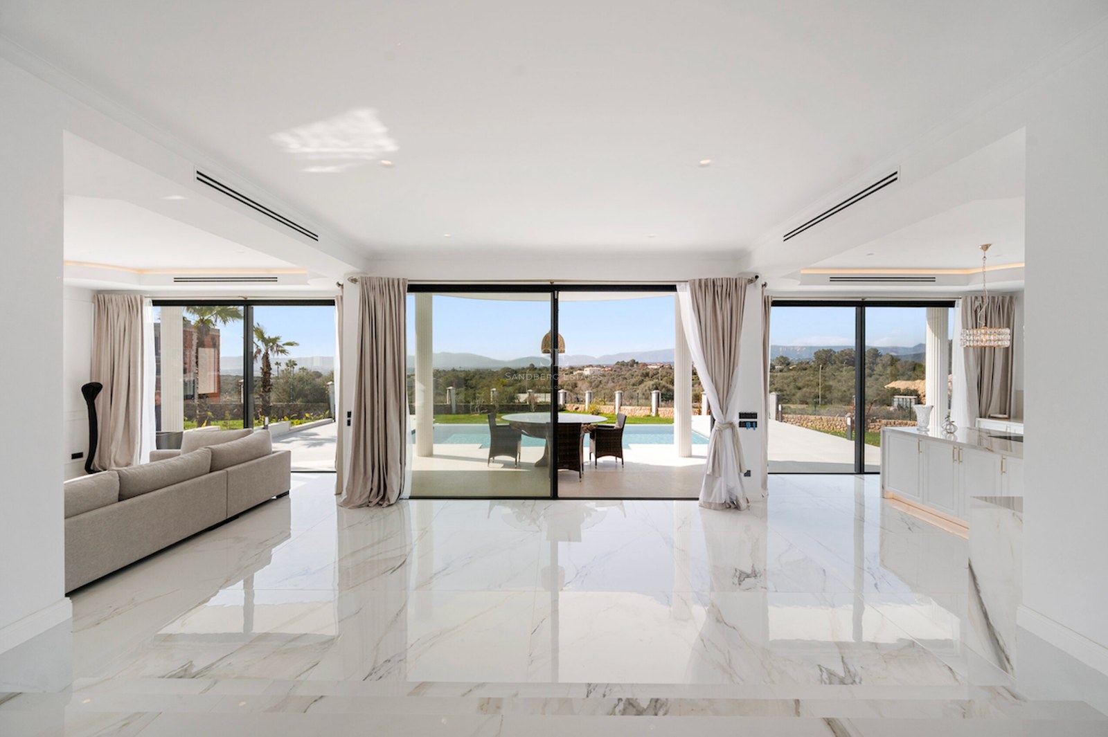 Salón luminoso con suelos de mármol blanco y ventanales de suelo a techo con vistas a la piscina y colinas.