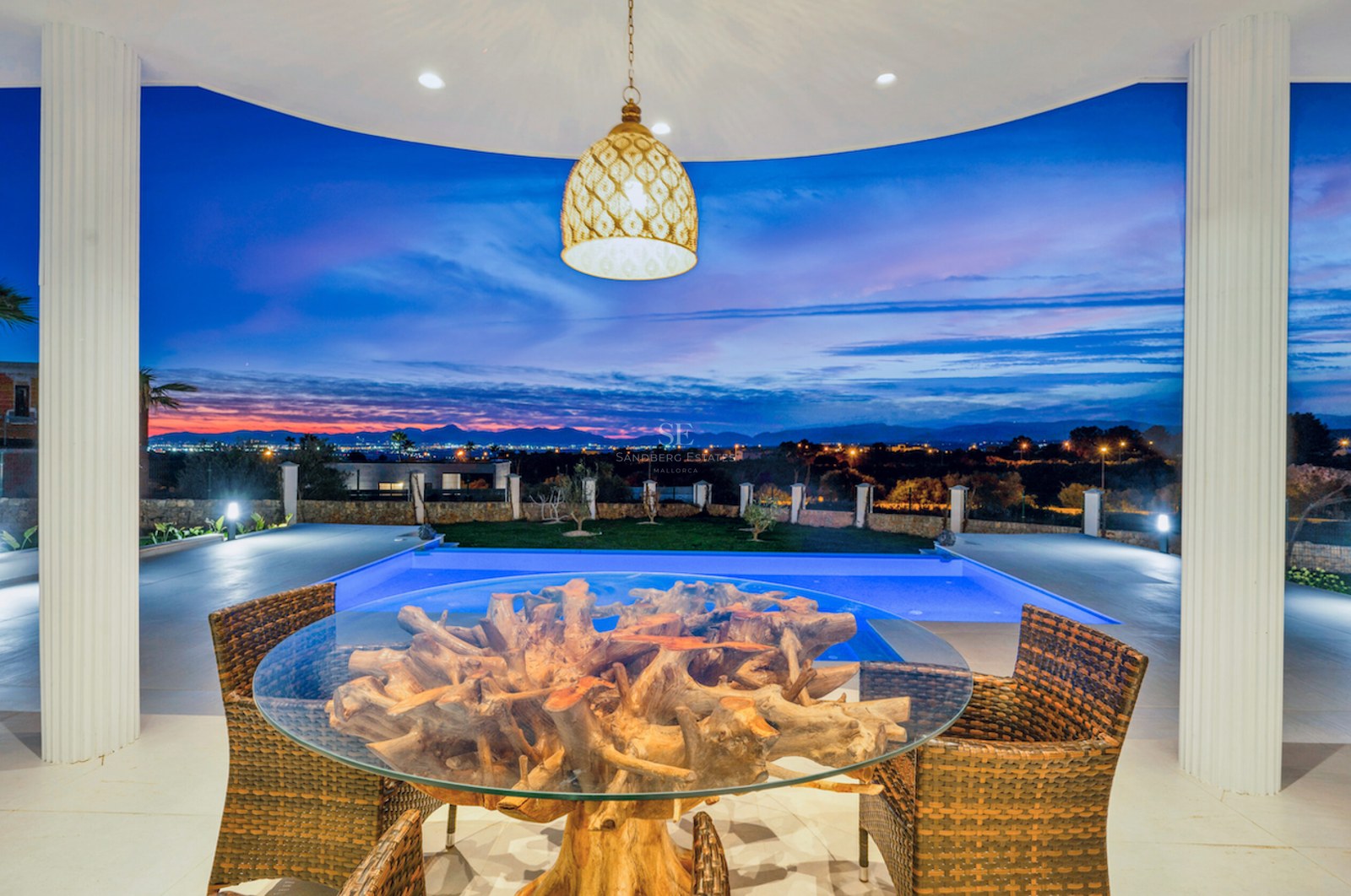 Terraza cubierta con mesa de raíz natural, sillas de mimbre y piscina iluminada con vistas lejanas al atardecer.