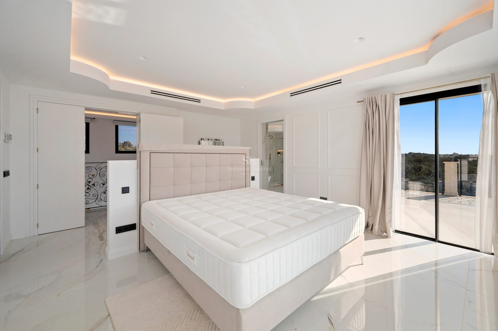 Dormitorio principal luminoso con suelos de mármol pulido, cabecero acolchado, iluminación indirecta y acceso a terraza.
