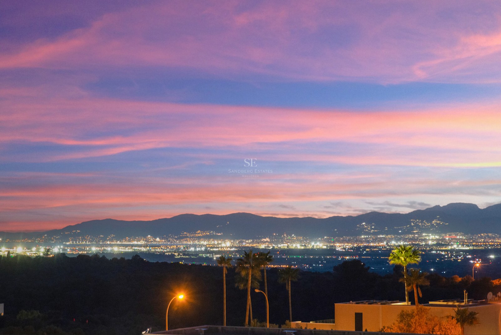 Atardecer con cielo rosado y violeta sobre montañas y luces de la ciudad con palmeras en primer plano.