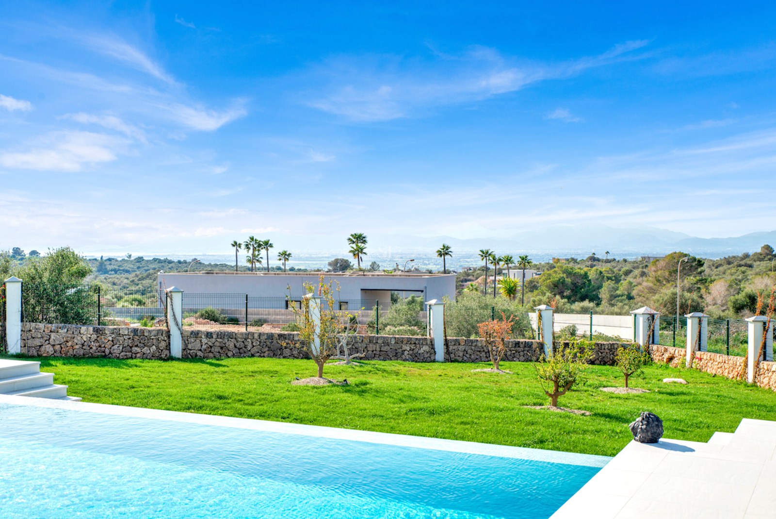 Piscina infinita turquesa y jardín con césped frente a un paisaje de montañas y cielo azul despejado.