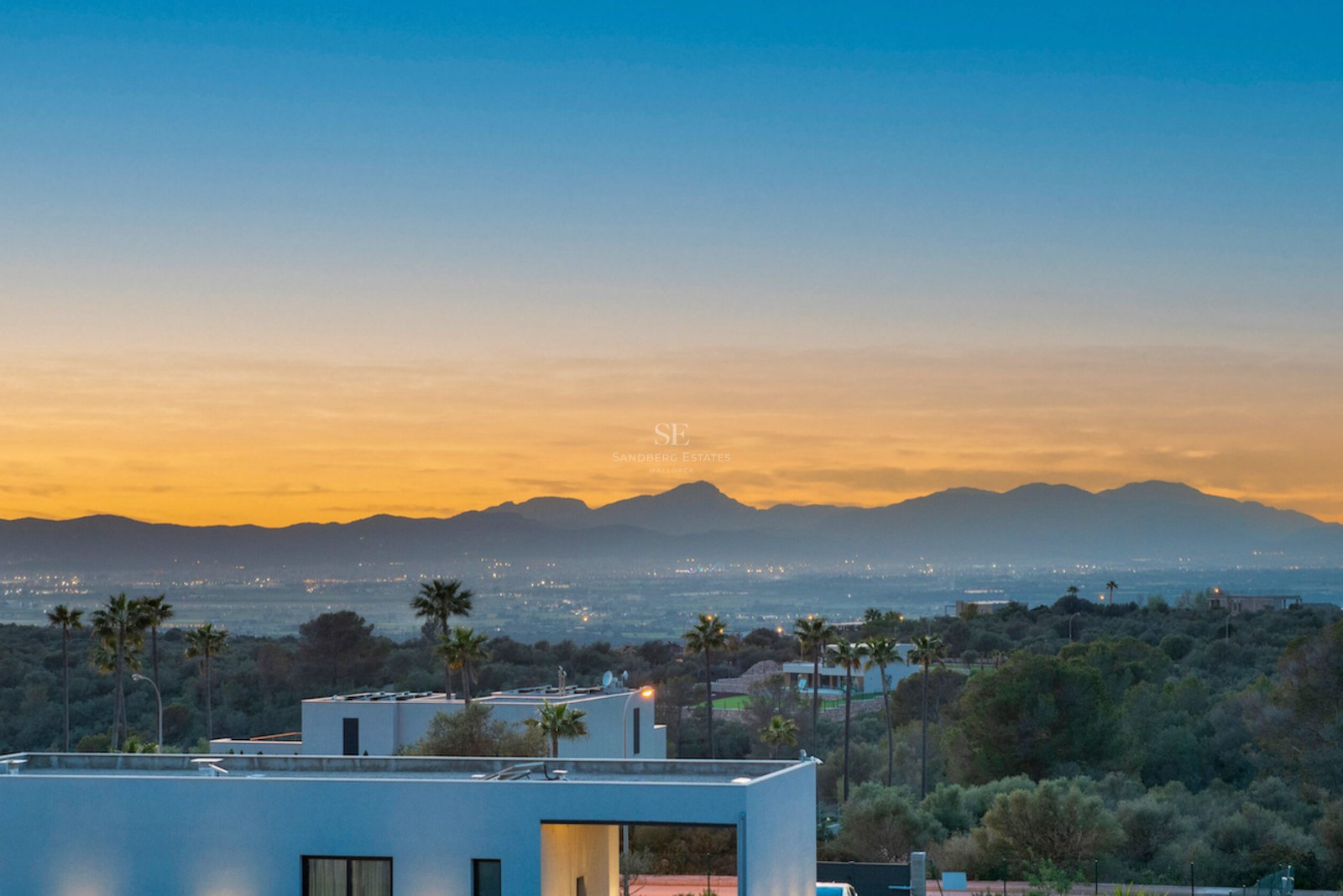 Villa moderna con vistas a un valle y montañas durante un atardecer dorado con luces de la ciudad al fondo.