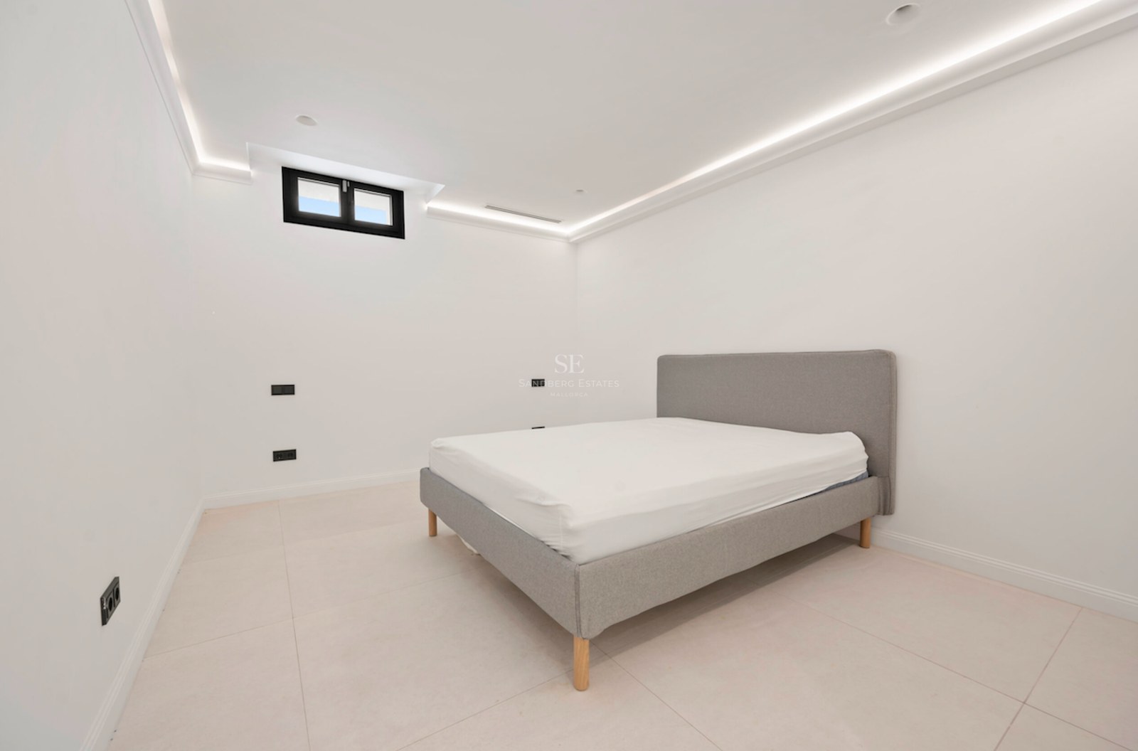 Habitación moderna con cama gris tapizada, suelos de gres claro y foseado de luz LED perimetral en el techo.