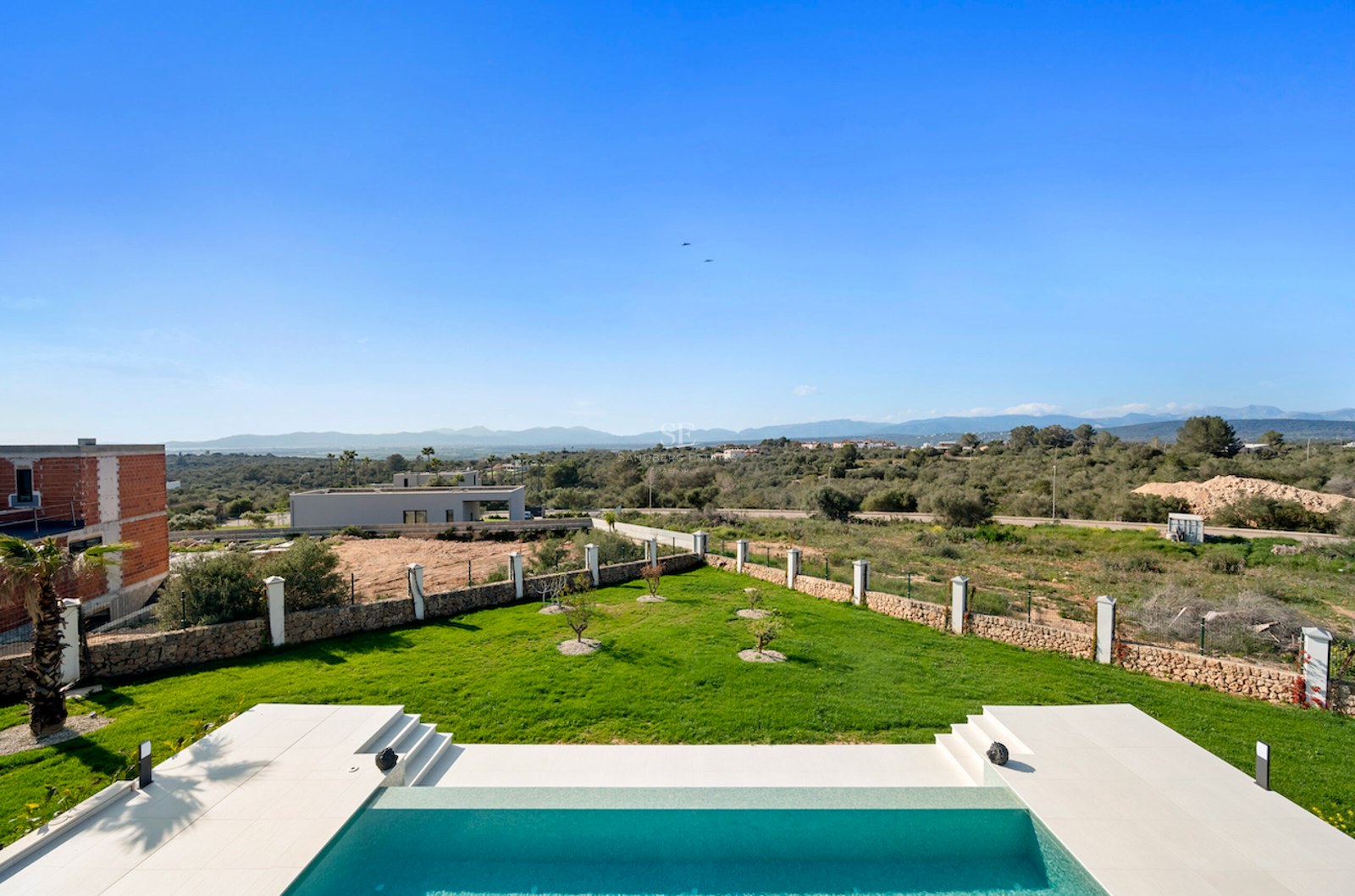 Piscina moderna y jardín de césped con muros de piedra y vistas panorámicas al mar y la montaña.