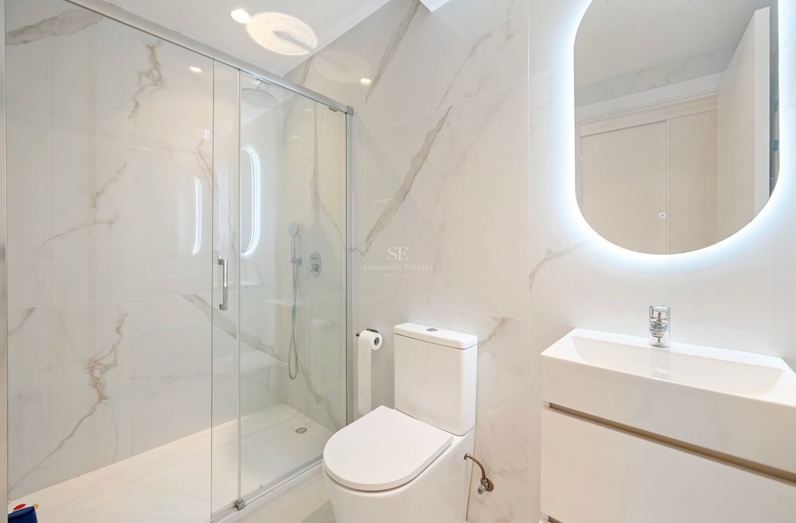 Baño moderno con paredes de efecto mármol, ducha de cristal y espejo ovalado retroiluminado sobre lavabo blanco.