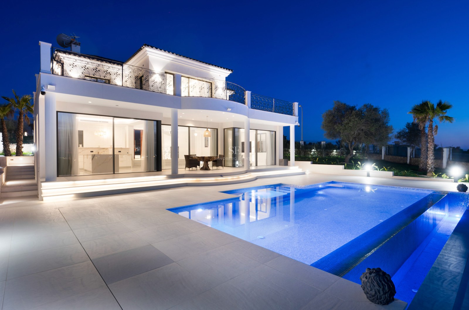 Villa de lujo blanca de dos plantas al atardecer con una gran piscina iluminada, terraza de piedra y balcones de forja.