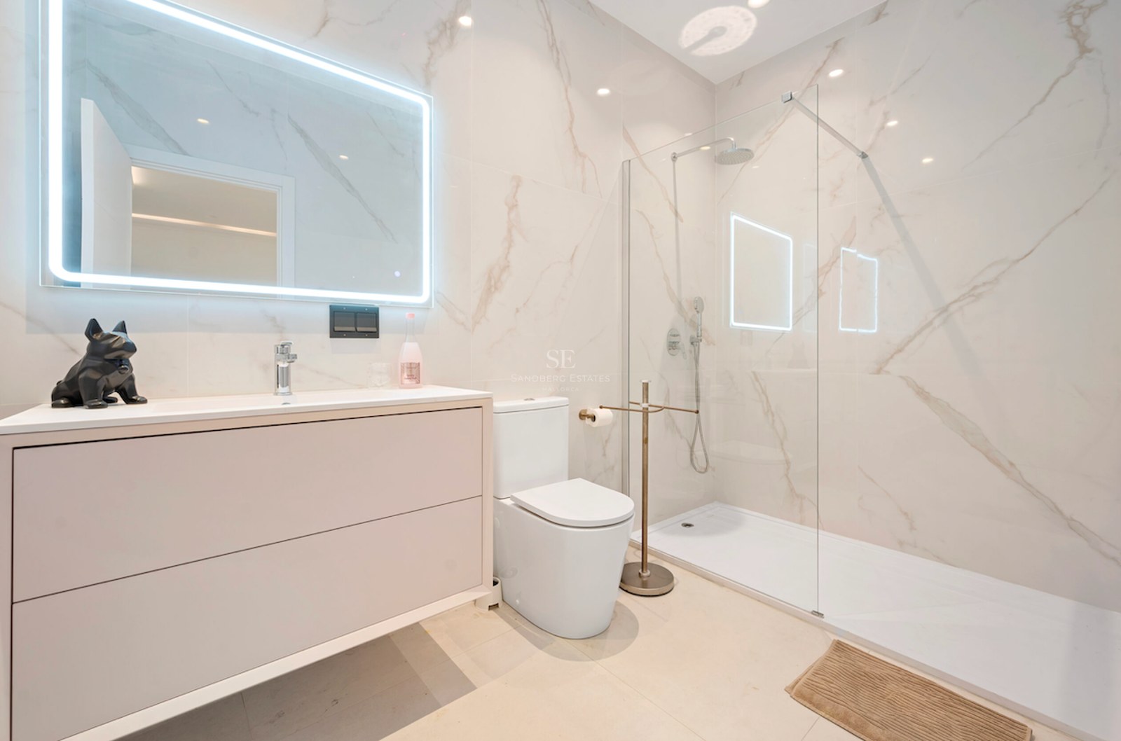 Baño luminoso con ducha de cristal, azulejos de mármol blanco, mueble de lavabo y espejo rectangular retroiluminado.