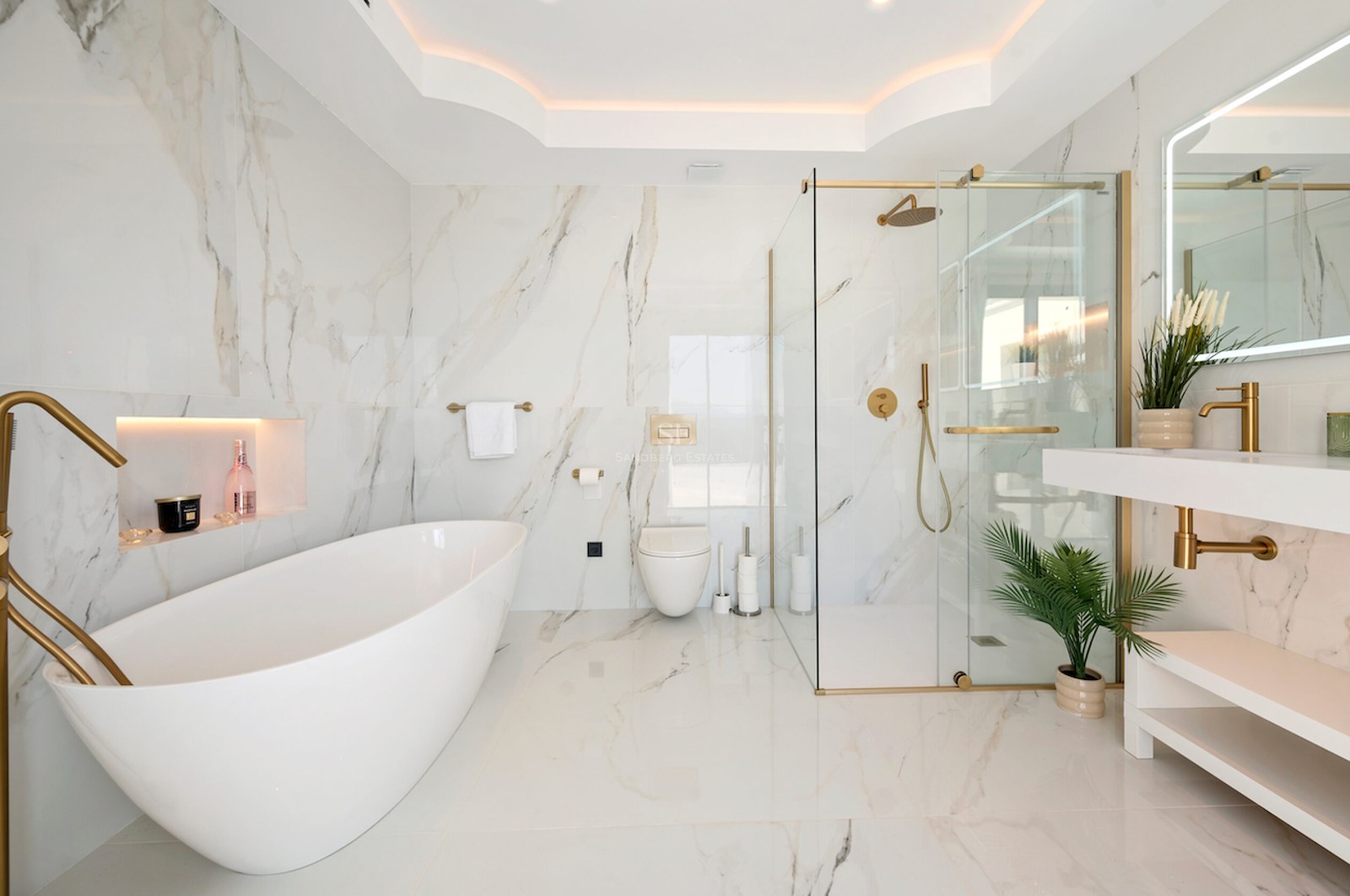 Baño de lujo con bañera exenta blanca, ducha de cristal y grifería dorada sobre paredes de mármol veteado.