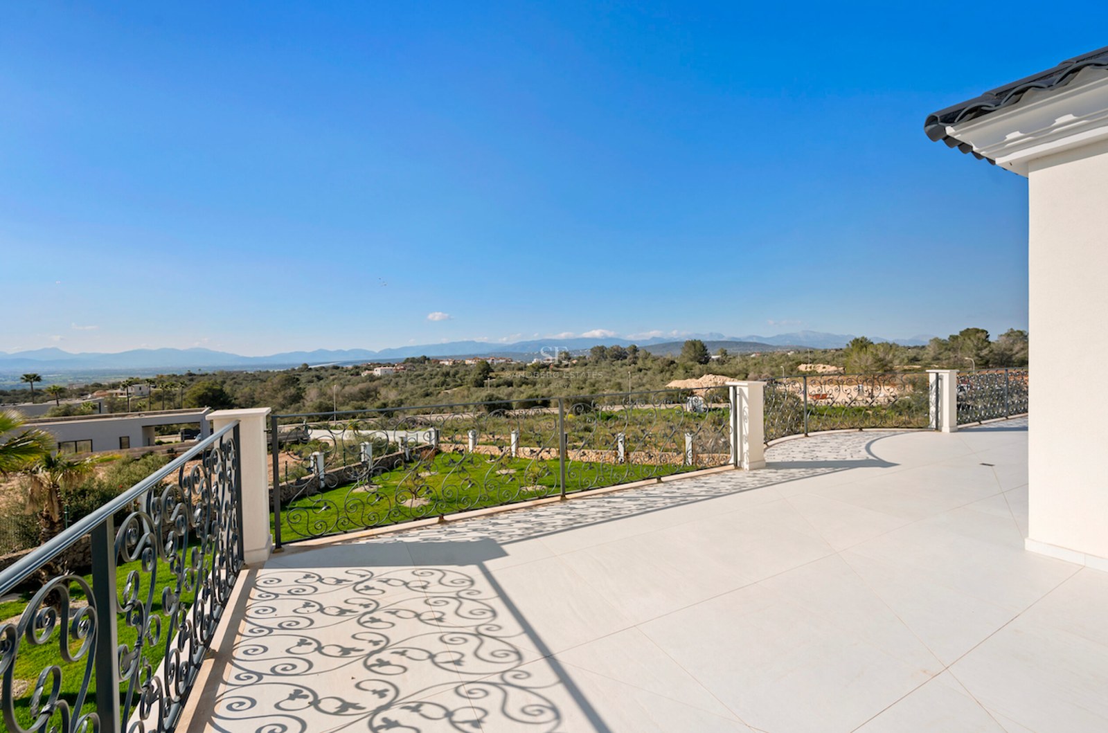Terraza con suelo color crema y barandilla de hierro forjado con vistas a un valle verde y montañas.