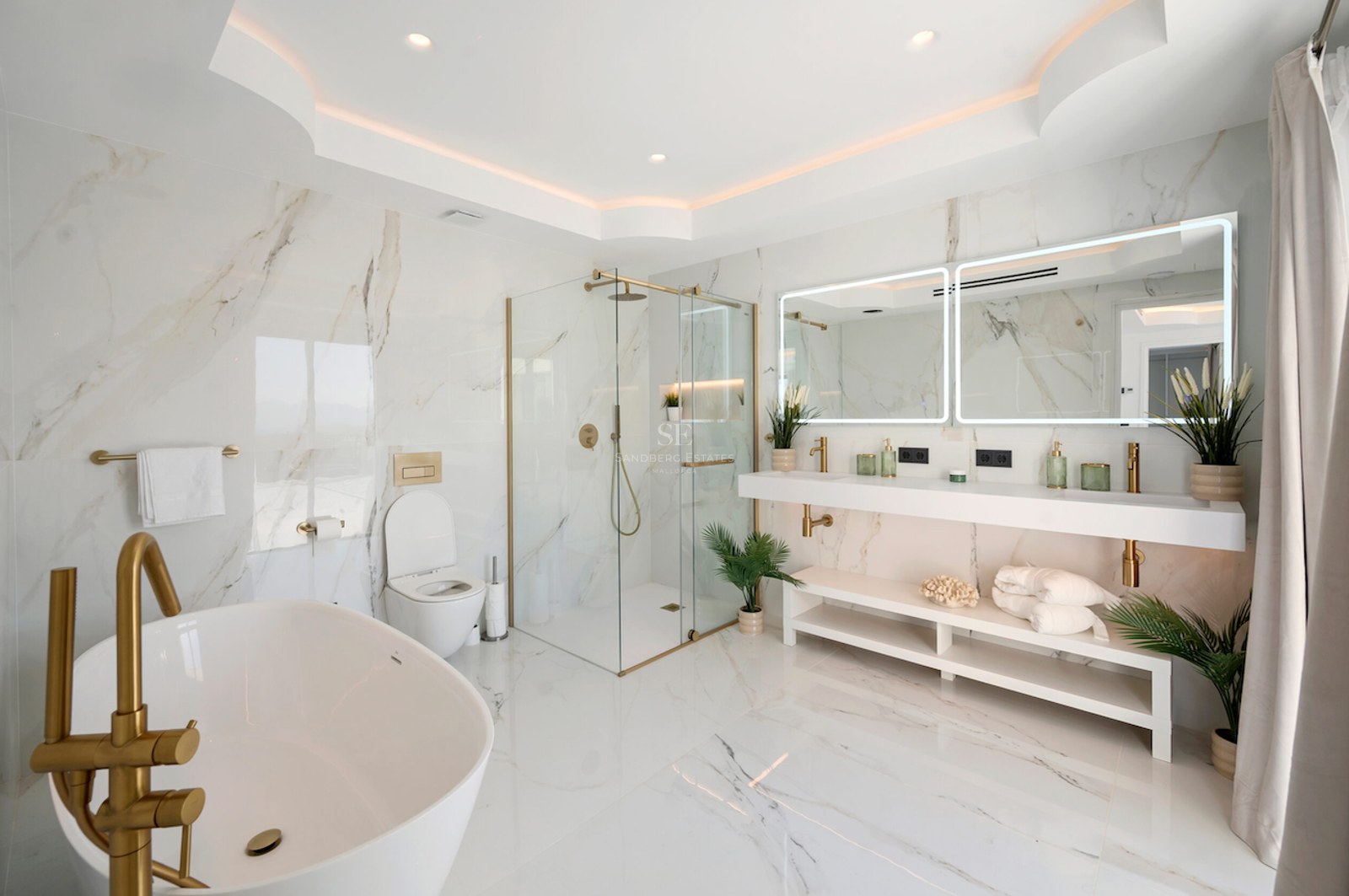 Baño de lujo con paredes de mármol, grifería dorada, bañera exenta, ducha de cristal y lavabo doble.