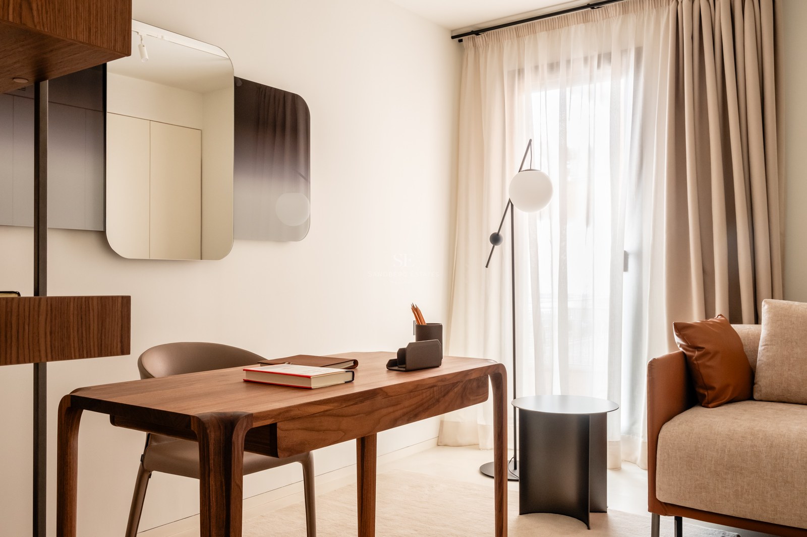 Modern wooden desk and leather chair in a bright room with sheer curtains and a minimalist floor lamp.