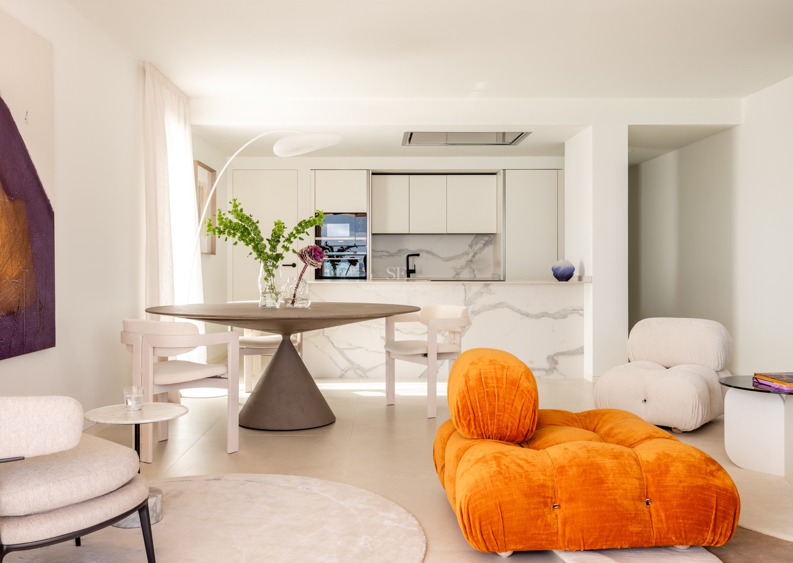 Bright open-plan living room and kitchen featuring an orange velvet sofa, marble backsplashes, and a round dining table.