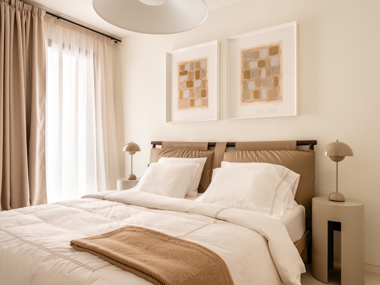 Modern bedroom with a tan leather headboard, white linens, and large windows with sheer curtains.