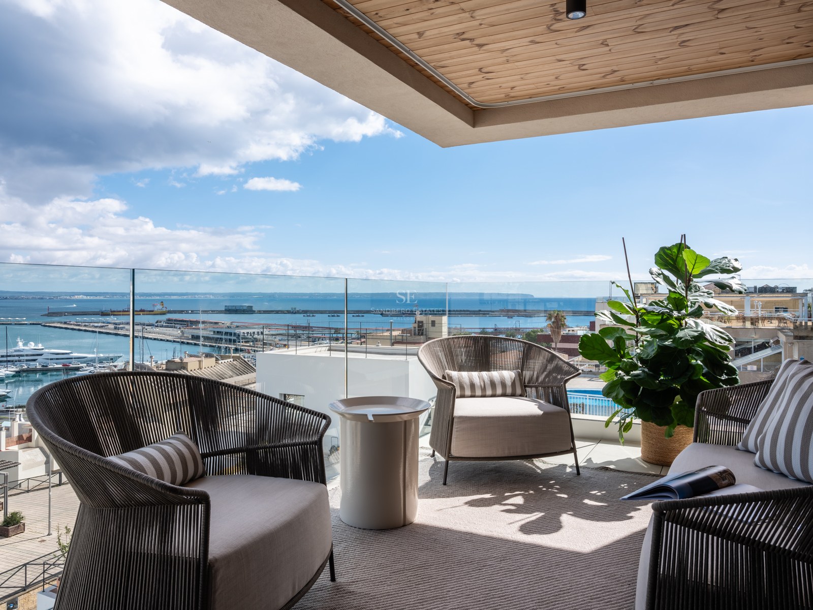 Contemporary terrace with grey woven armchairs, glass railing, and a view of a Mediterranean harbor with yachts.