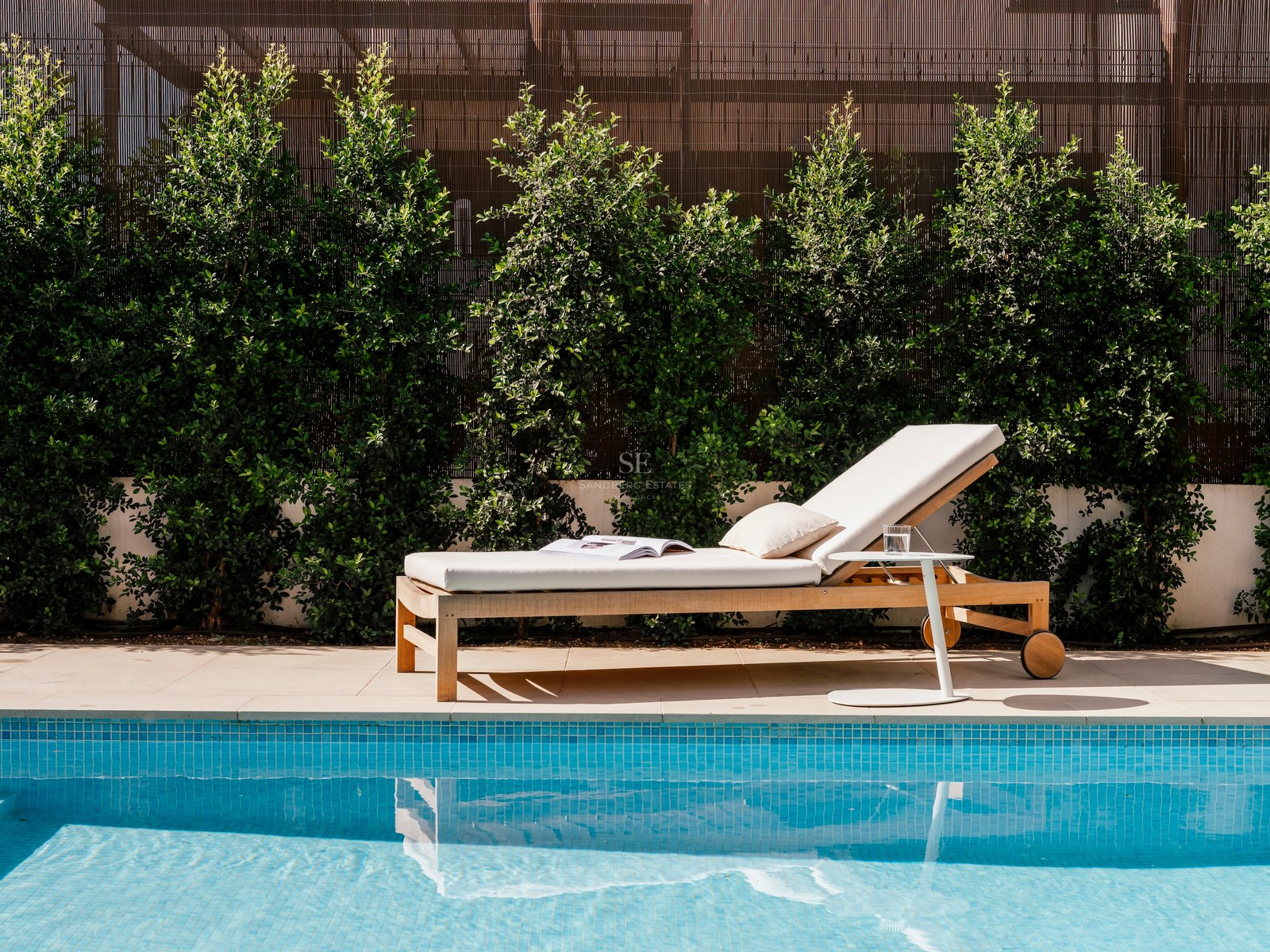 Bain de soleil en bois avec coussins blancs à côté d'une piscine en mosaïque bleue et d'une haie verdoyante.