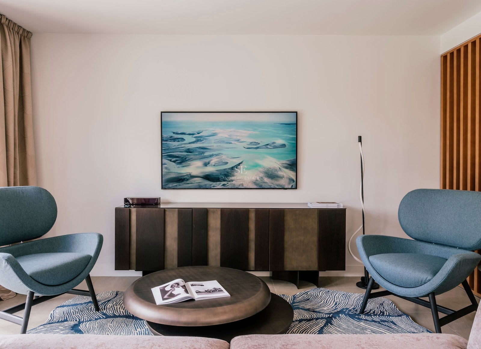 Deux fauteuils bleu canard face à une table basse en bronze et un buffet en bois sombre dans une pièce lumineuse.