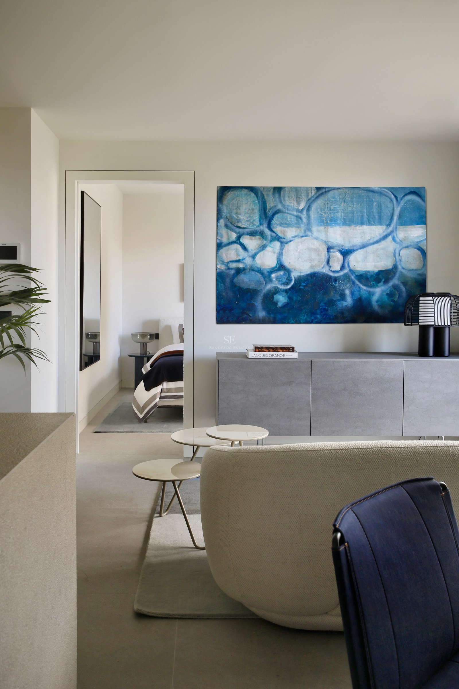 Modern living room with a large blue abstract painting over a grey sideboard, looking into a minimalist bedroom.