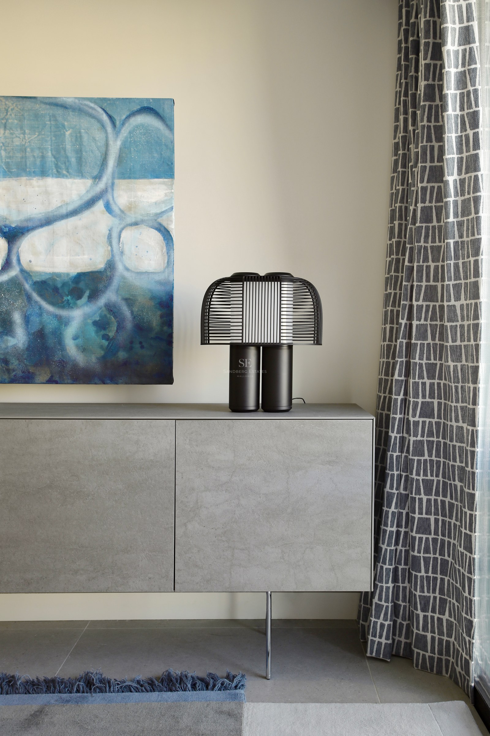 A grey stone-finish sideboard with a black designer lamp, an abstract blue painting, and patterned curtains.
