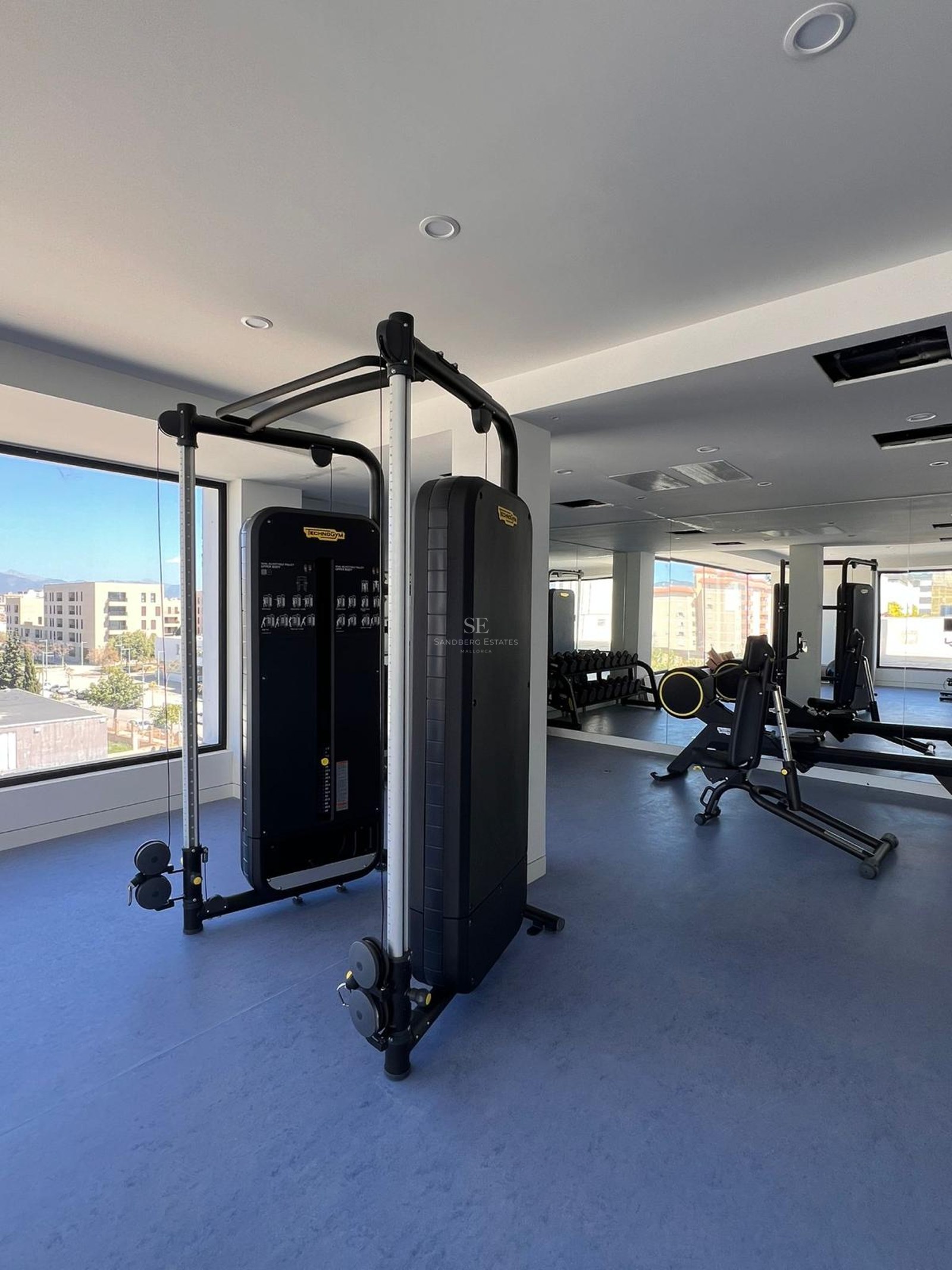 Modern home gym featuring black Technogym equipment, blue rubber flooring, and large windows with urban views.