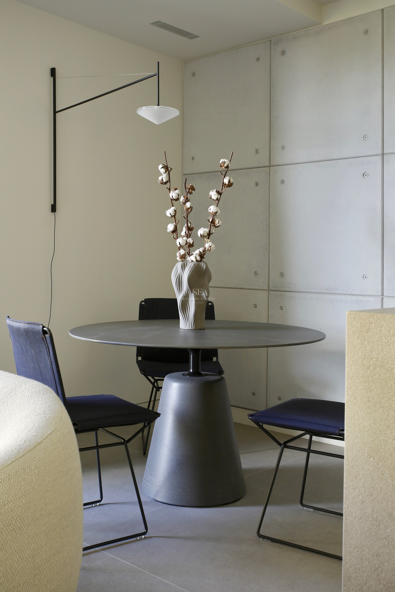 Modern round grey pedestal table with navy chairs against a concrete panel wall and minimalist wall lamp.
