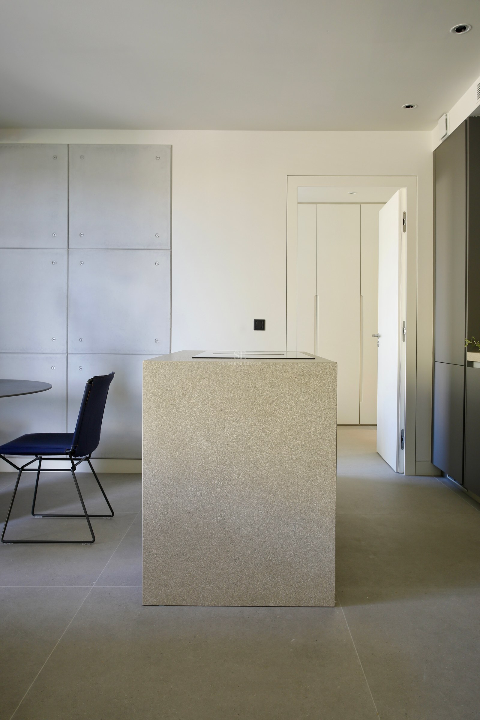 Modern kitchen featuring a textured stone island, concrete wall panels, and a navy blue chair on grey tiled floors.