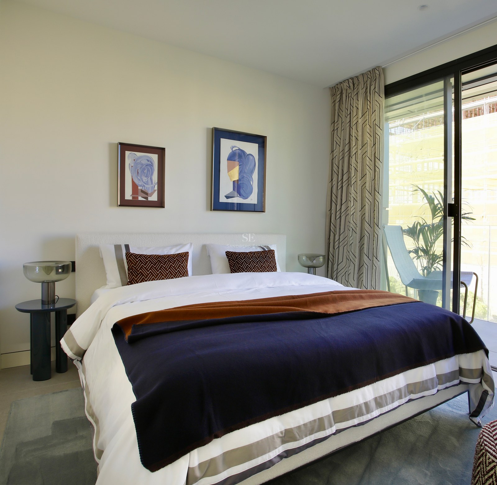 Contemporary bedroom featuring white bedding, navy throw, and floor-to-ceiling glass doors opening to a sunlit balcony.