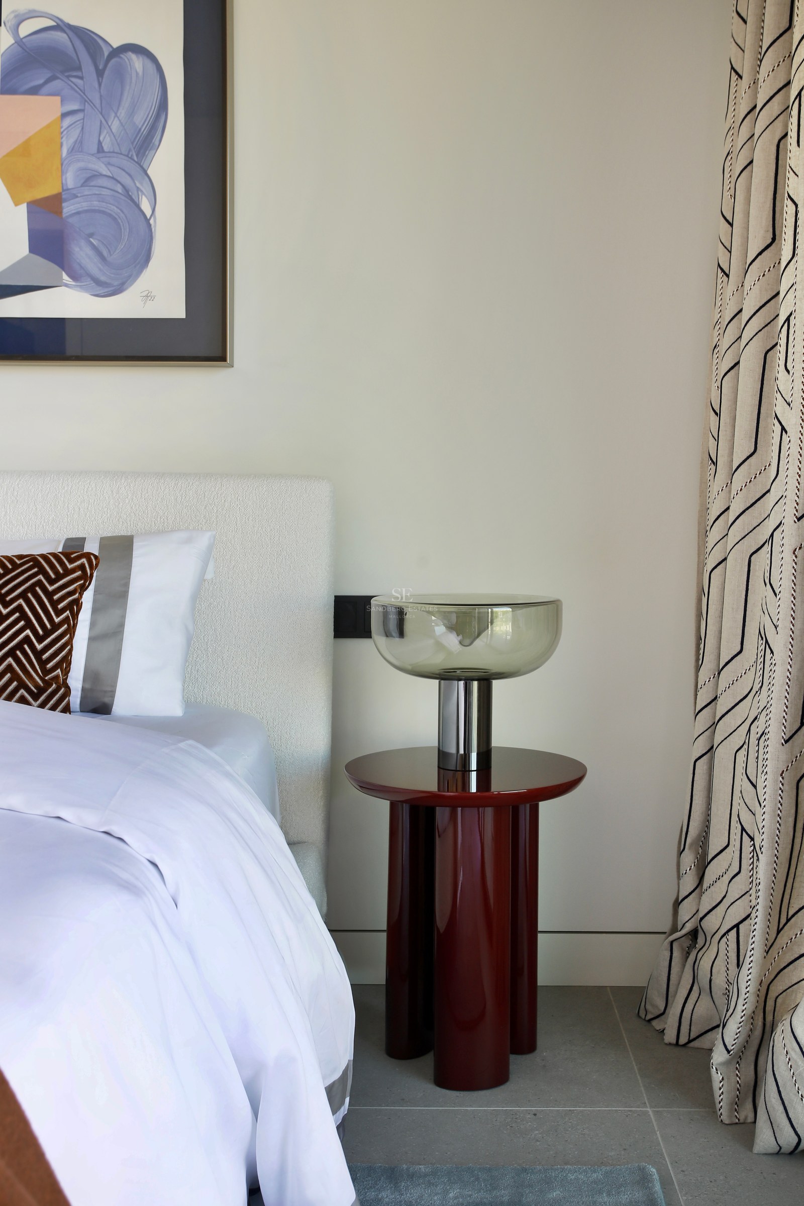 Bedroom detail featuring a white bouclé headboard, red lacquered side table, glass lamp, and abstract art.