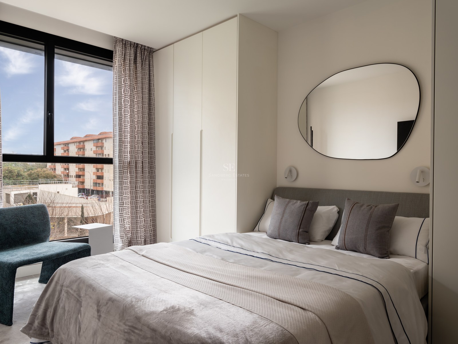 Modern bedroom featuring a white bed, organic mirror, built-in wardrobes, and a blue armchair by a large window.