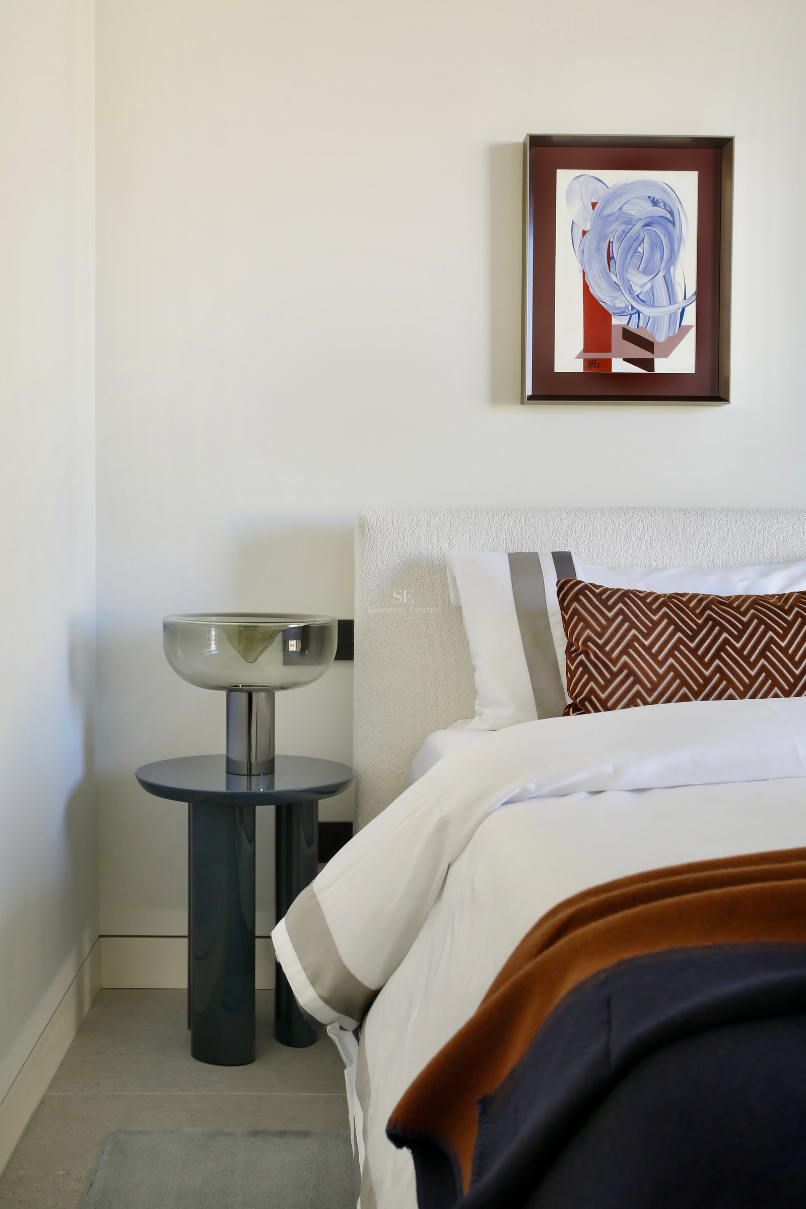 Close-up of a white bouclé headboard, designer glass lamp on a blue side table, and abstract wall art.