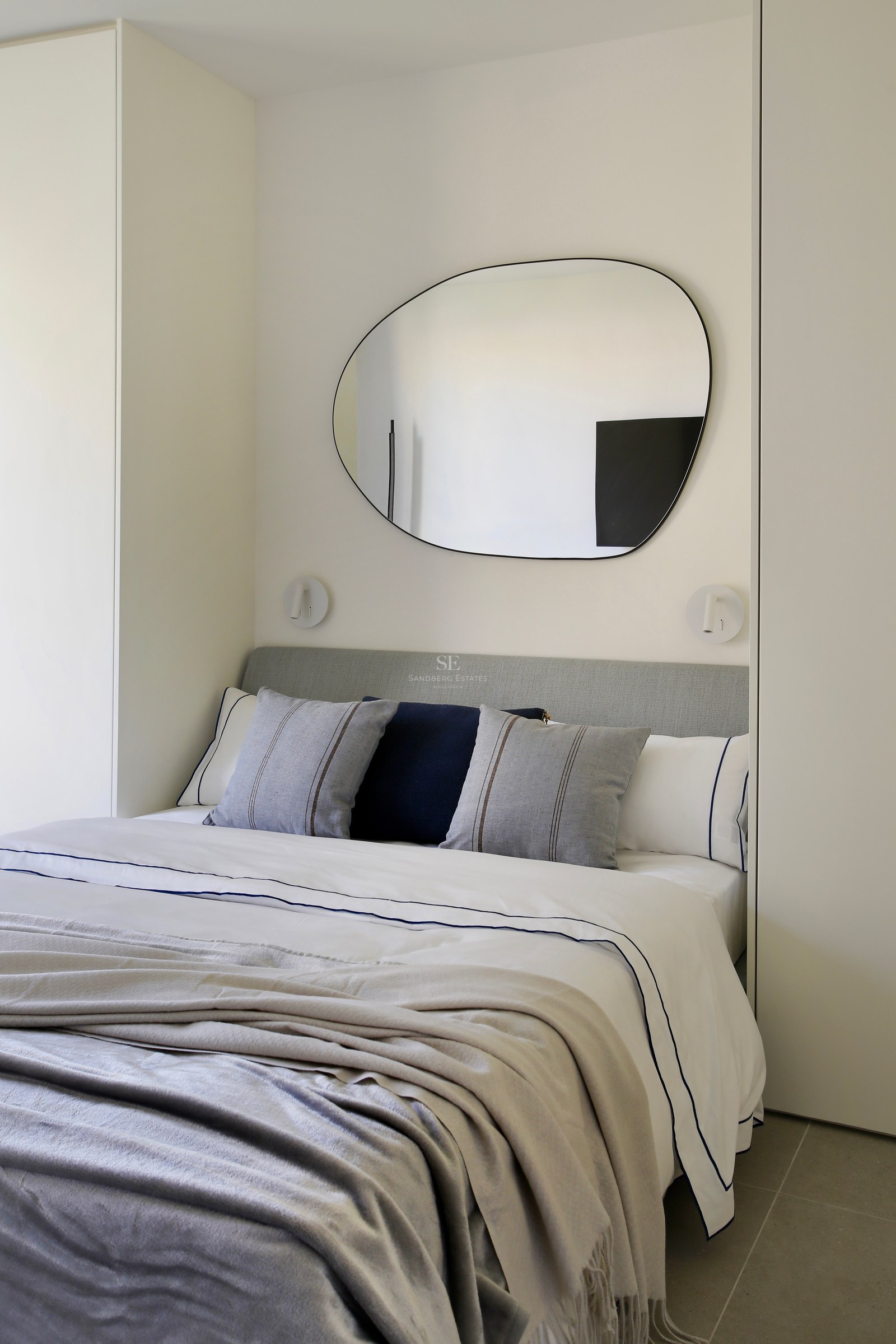 A modern bedroom featuring a neutral-toned bed, an organic-shaped wall mirror, and integrated white wardrobes.