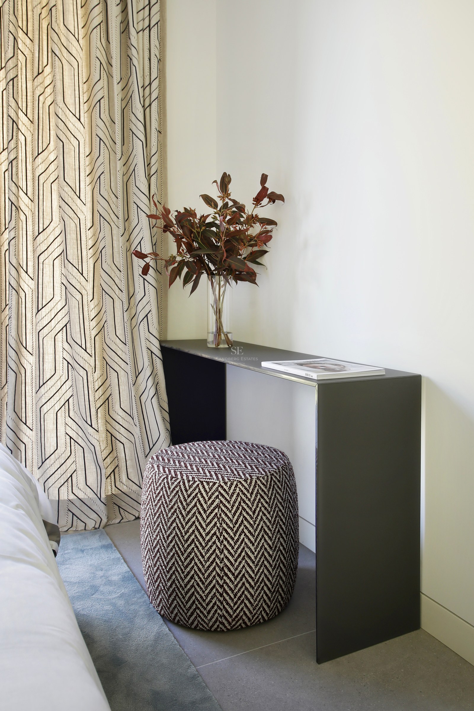 Modern bedroom corner featuring a minimalist gray console, patterned ottoman, and geometric curtains with natural light.