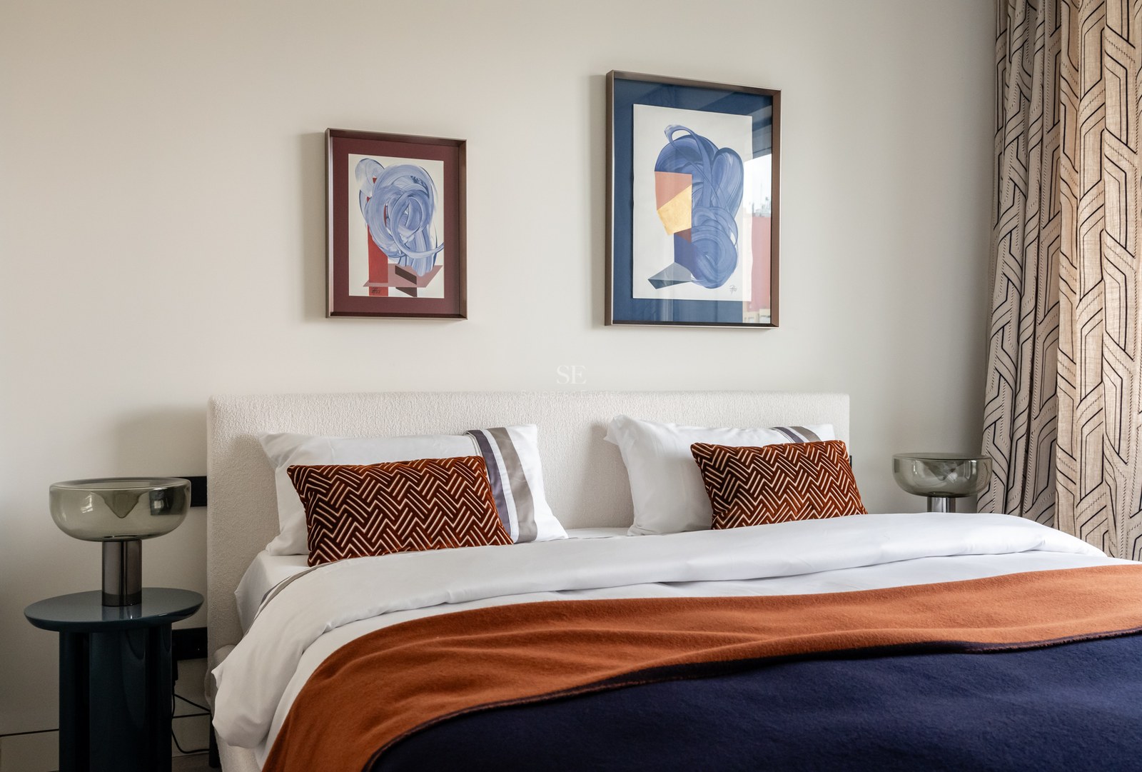 Modern bedroom featuring a white bouclé headboard, navy and terracotta bedding, and abstract artwork above the bed.