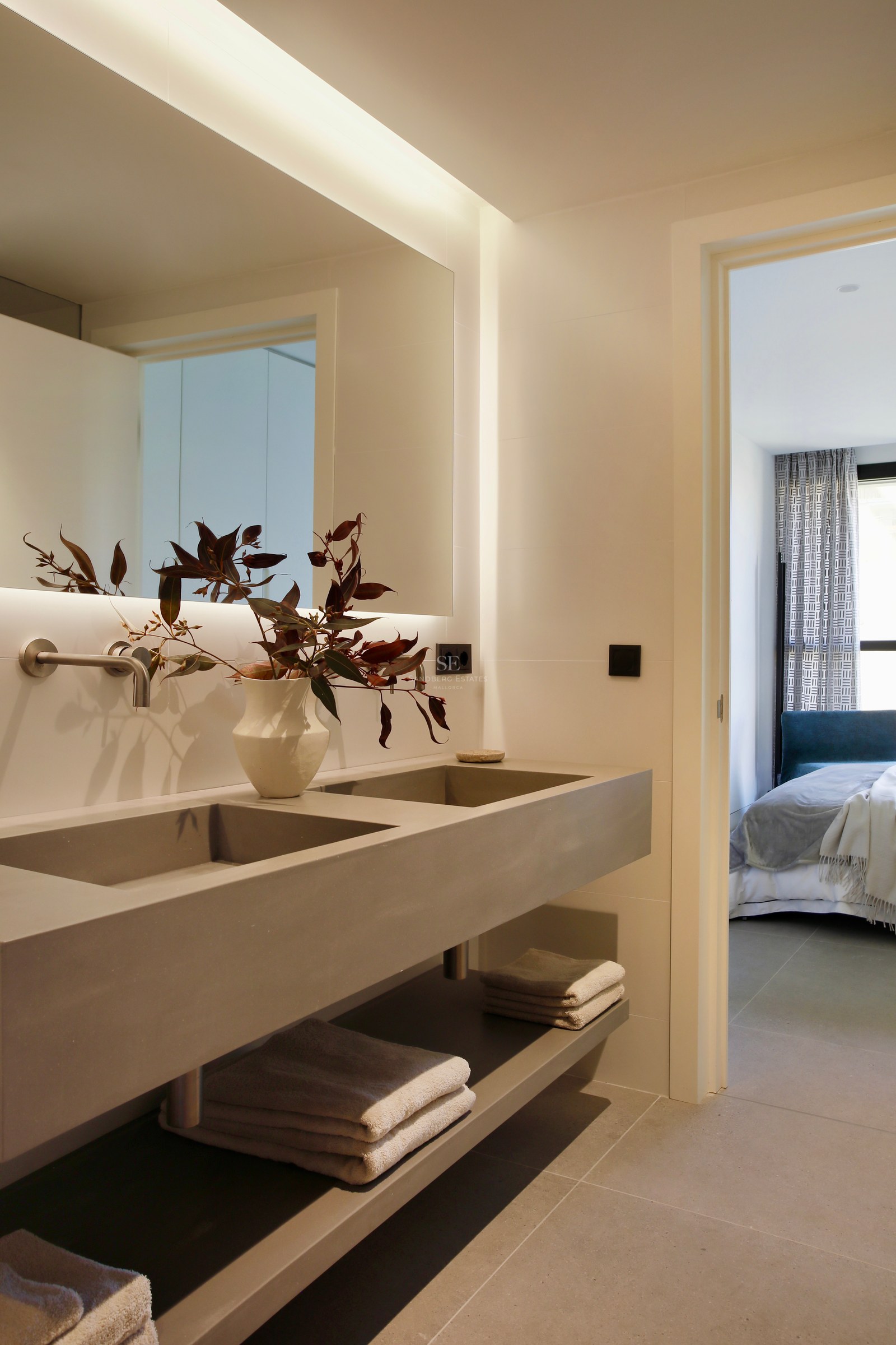 Contemporary bathroom with a grey double sink, large backlit mirror, and view into the adjacent bedroom.