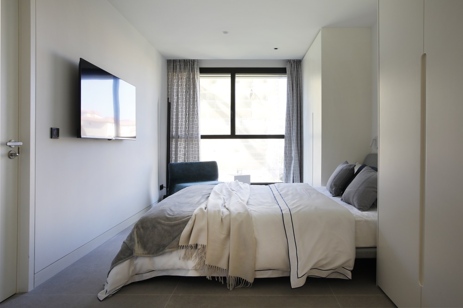 Minimalist bedroom with white walls, a large window, wall-mounted TV, and grey tiled flooring.