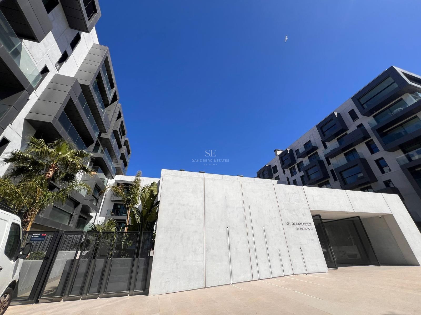 Contemporary apartment building with concrete facade, glass balconies, and palm trees under a clear blue sky.