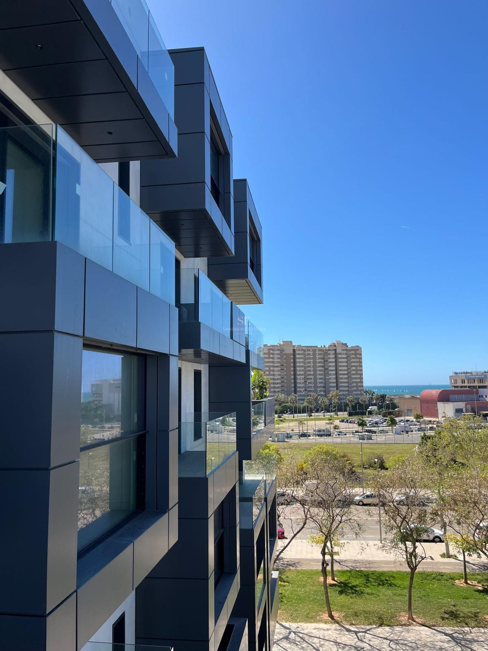 Modern dark grey modular facade with glass balconies overlooking an urban park and the Mediterranean Sea.