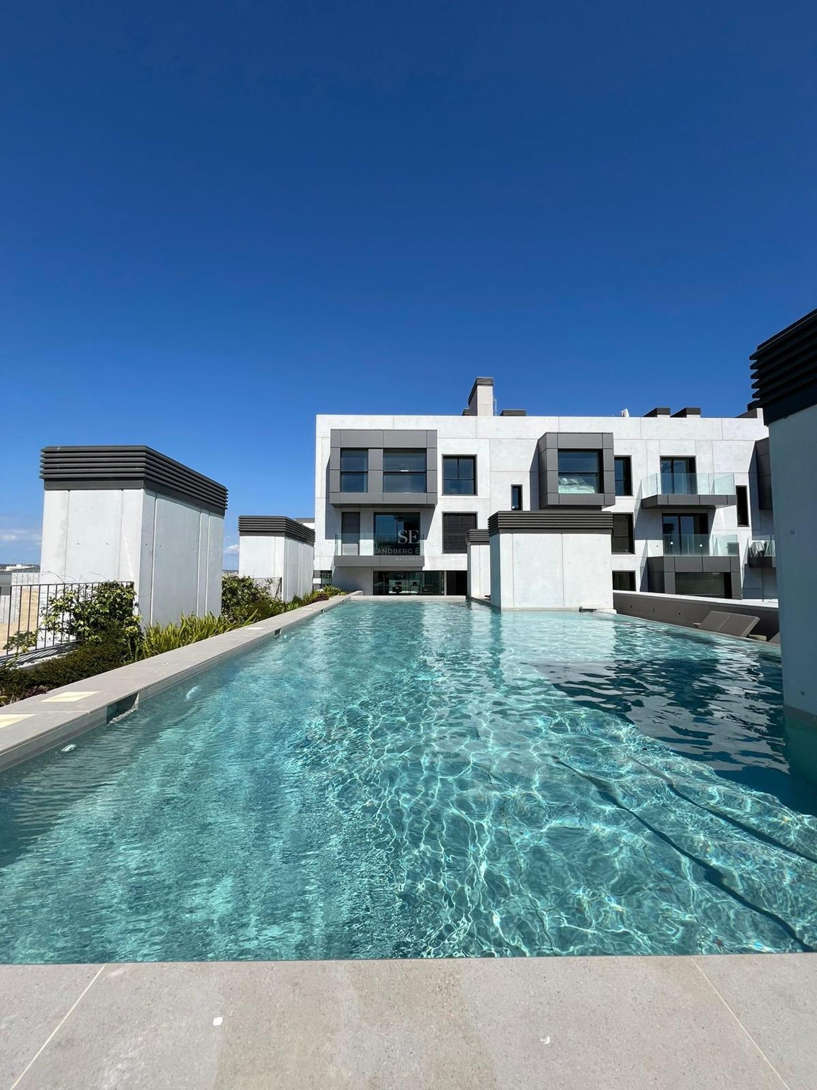 A long turquoise rooftop swimming pool with stone edges in front of a white and grey contemporary apartment building.