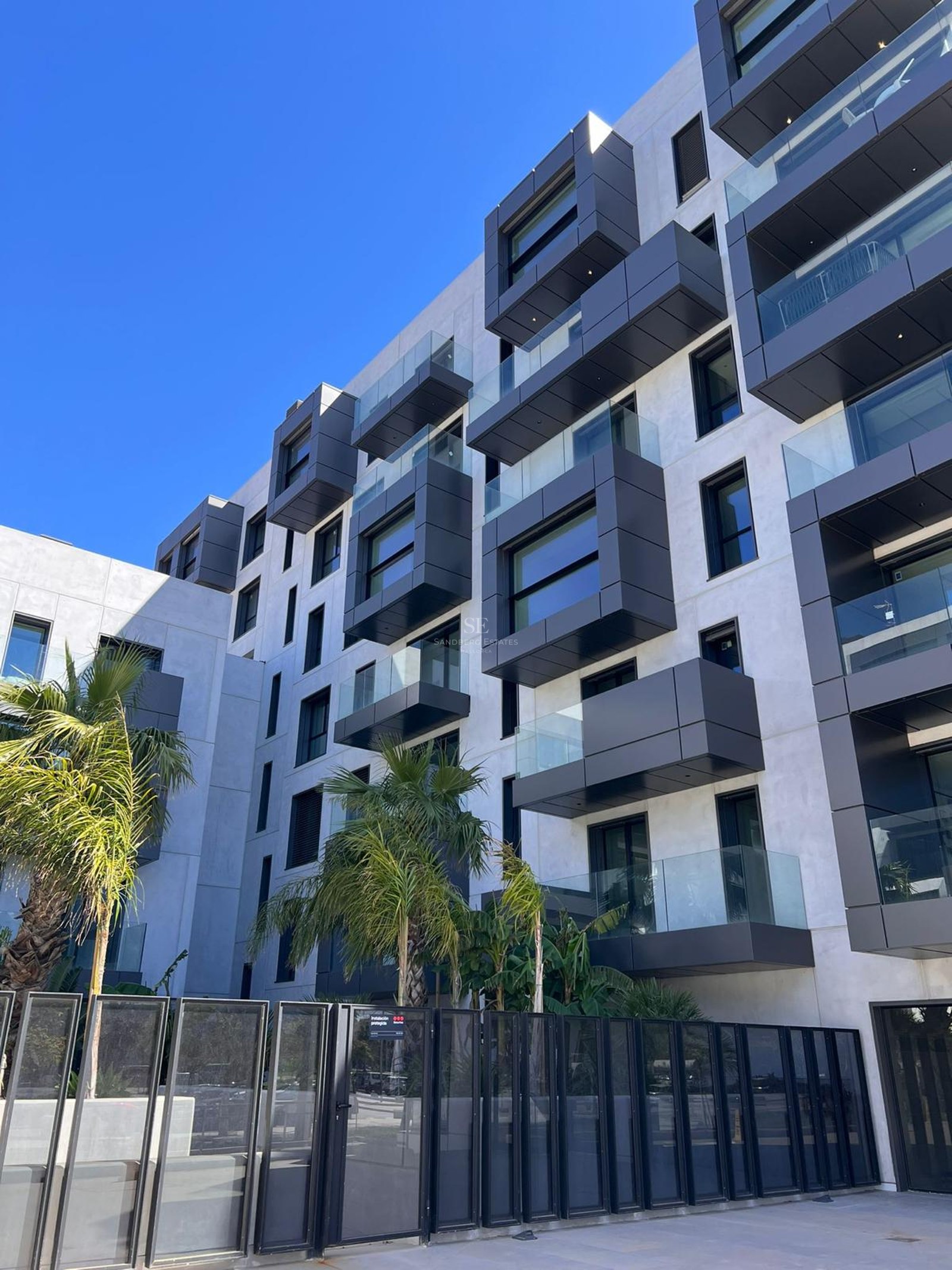 Modern multi-story apartment building with off-white walls, dark gray boxy balconies, glass railings, and palm trees.