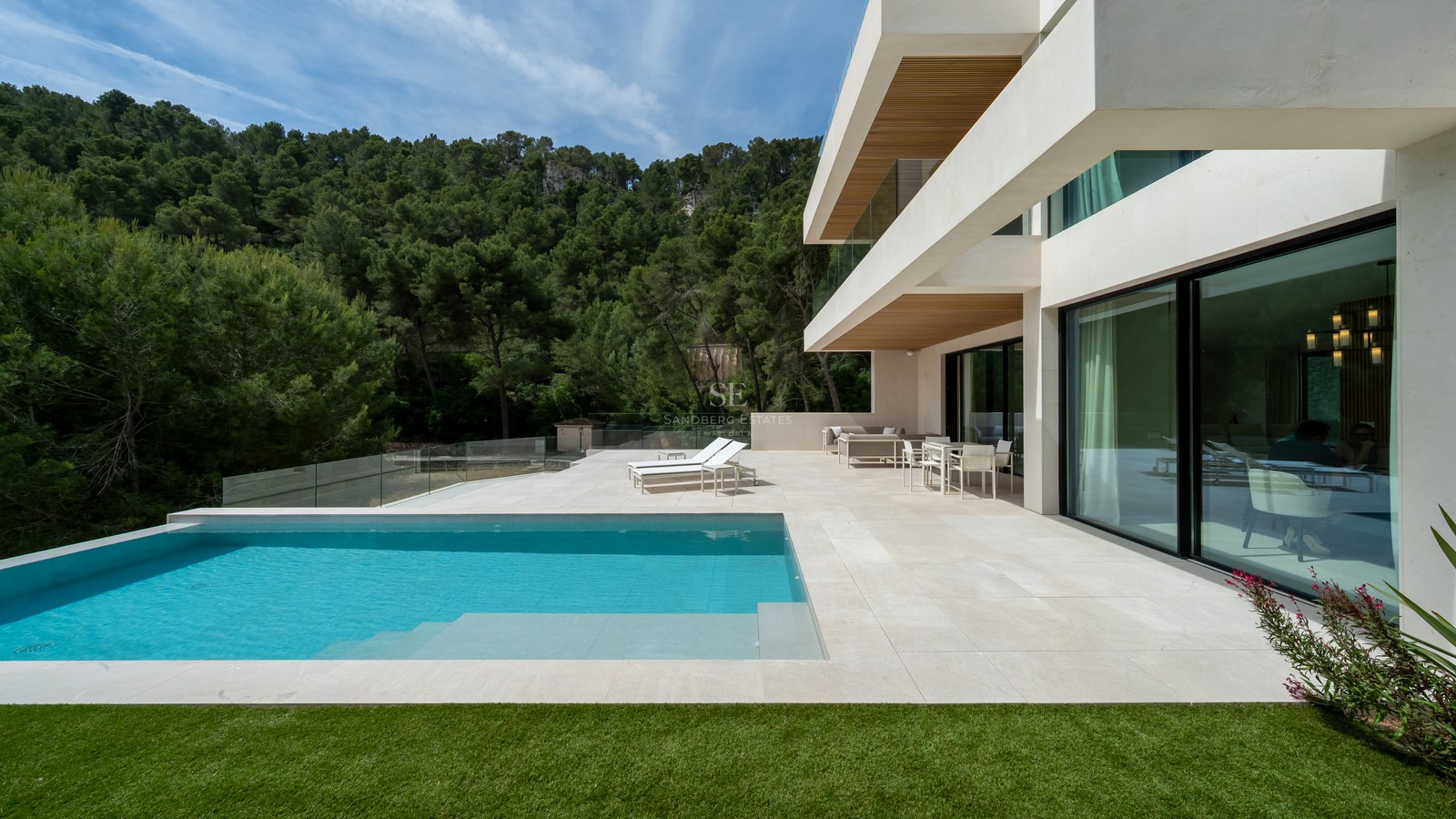 Villa blanca moderna con piscina turquesa, terraza de piedra y barandillas de cristal frente a un bosque de pinos.