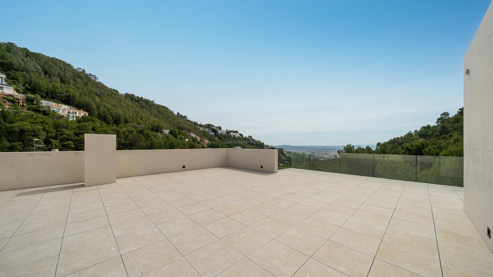 Amplia terraza de piedra natural con barandilla de cristal y vistas a colinas verdes y cielo despejado.